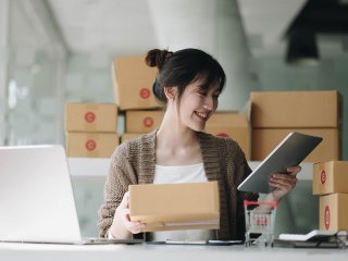 Eine lächelnde Frau verpackt verschiedene Pakete, während sie auf ein Tablet, blickt